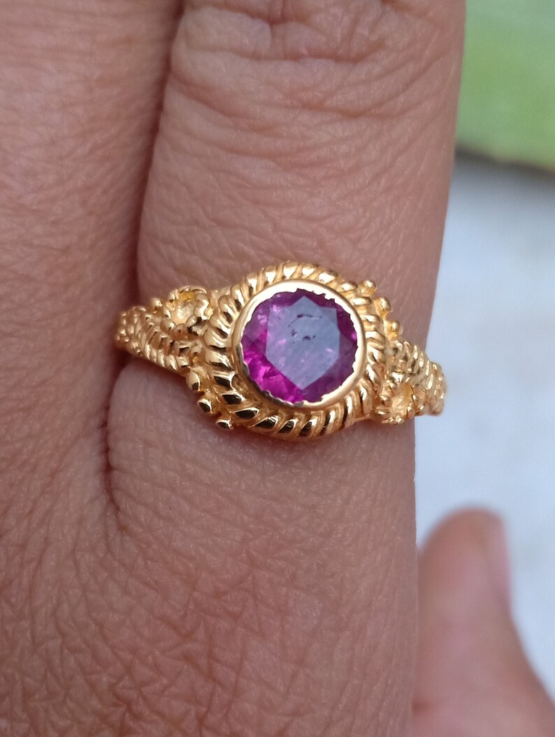 Pode incluir: Um anel de ouro com uma grande pedra preciosa magenta redonda. O anel tem uma banda detalhada e texturizada com desenhos florais e semelhantes a cordas. A pedra &eacute; definida em uma configura&ccedil;&atilde;o de moldura.