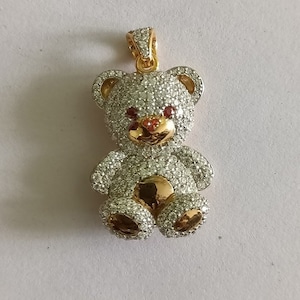 Pode incluir: Um pingente de urso de pelúcia dourado com pedras de zircônia cúbica transparentes e vermelhas. O pingente tem um laço na parte superior para prender a uma corrente.