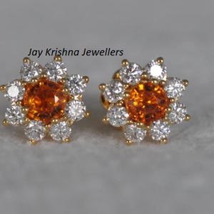 May include: A pair of flower-shaped stud earrings. Each earring has a central orange gemstone surrounded by white gemstones, set in gold-coloured metal. The earrings are displayed on a grey surface.