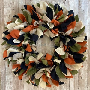 May include: A fall-themed wreath made of felt fabric in shades of orange, green, black, and cream. The wreath is made of many individual felt pieces, creating a full and fluffy look.