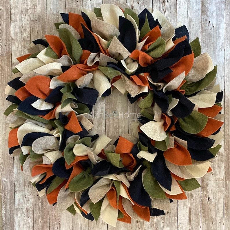 May include: A fall-themed wreath made of felt fabric in shades of orange, green, black, and cream. The wreath is made of many individual felt pieces, creating a full and fluffy look.