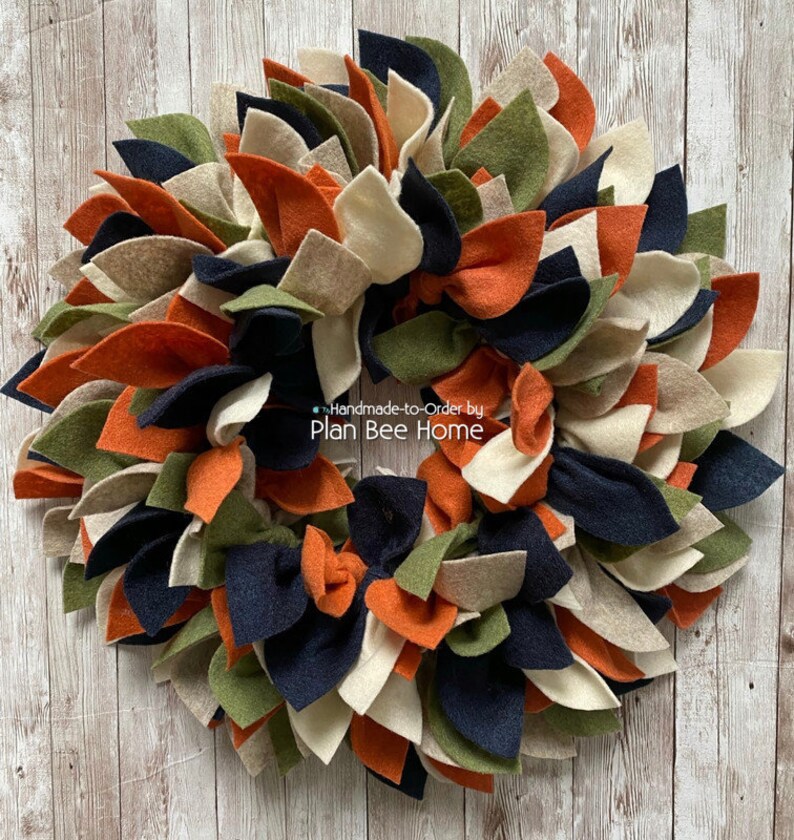 May include: A fall-themed wreath made of felt in shades of orange, green, cream, and navy blue. The wreath is handmade and features a variety of leaf shapes. The text "Handmade-to-Order by Plan Bee Home" is visible on the wreath.