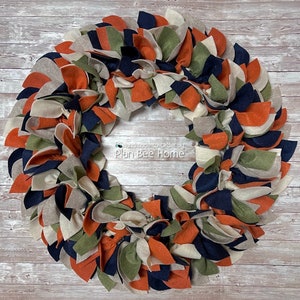 May include: A handmade felt wreath in shades of orange, beige, green, and navy blue. The wreath is made of multiple layers of felt, creating a textured and layered look. The wreath is handmade to order by Plan Bee Home.