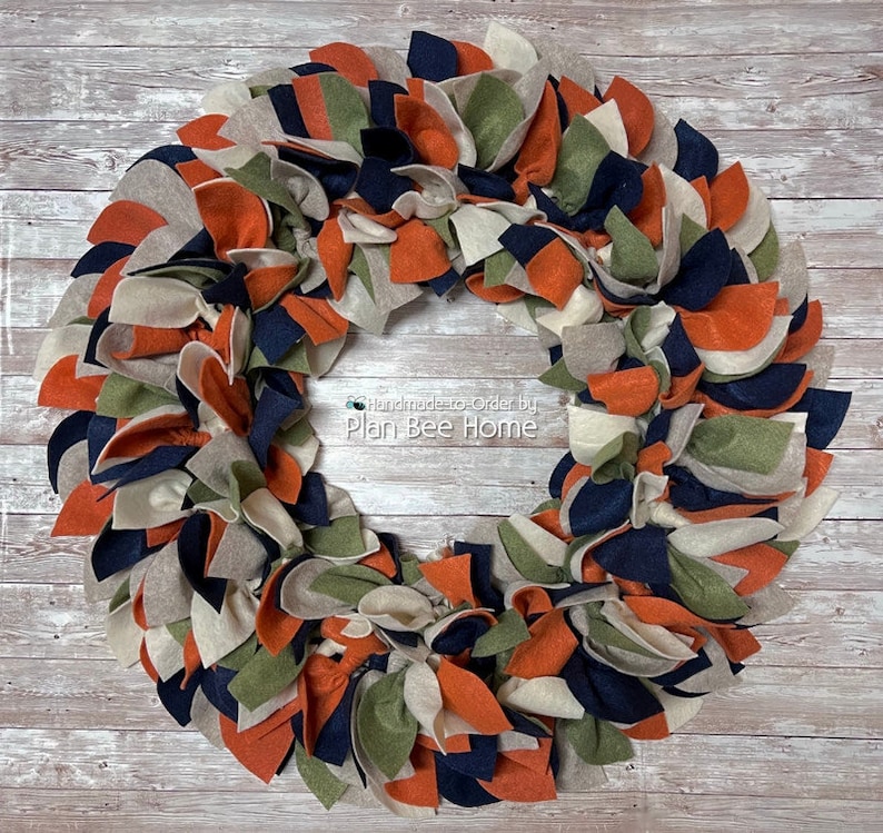 May include: A handmade felt wreath in shades of orange, beige, green, and navy blue. The wreath is made of multiple layers of felt, creating a textured and layered look. The wreath is handmade to order by Plan Bee Home.