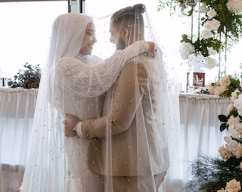 May include: A bride and groom embrace under a white veil with pearl accents. The couple is standing in a room with white curtains and a floral arrangement in the background.