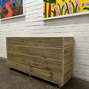 May include: A wooden slatted storage cabinet with a light brown finish. The cabinet is standing in front of a white brick wall.