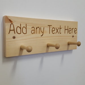 Op de afbeelding: Een houten kapstok met drie haken. De kapstok is gemaakt van natuurlijk hout en heeft de tekst "Add any Text Here" gegraveerd op de voorkant.