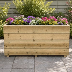 May include: A rectangular wooden planter box constructed from horizontal grooved planks with a natural light brown finish. The planter is filled with a variety of vibrant flowers in shades of purple, pink, white, and yellow, supported by small feet.