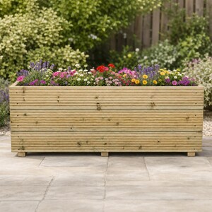 May include: A rectangular wooden planter box with horizontal slats, featuring a natural light brown wood grain finish. The planter is filled with a variety of colourful flowers, including purple lavender, bright red, pink, and yellow blossoms.