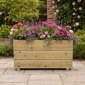 May include: A rectangular wooden planter box constructed from horizontal planks with a natural finish, filled with a vibrant arrangement of pink, purple, and white flowers, including lavender and petunias, supported by small block feet.