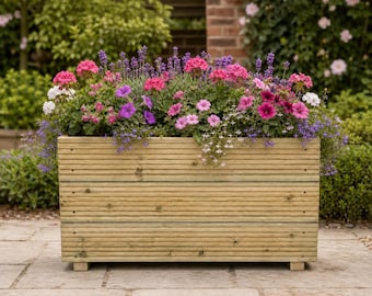May include: A rectangular wooden planter box constructed from horizontal planks with a natural finish, filled with a vibrant arrangement of pink, purple, and white flowers, including lavender and petunias, supported by small block feet.