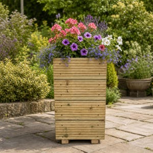 May include: A rectangular wooden planter box with horizontal slats and small feet, filled with a vibrant arrangement of pink, purple, blue, and white flowers, including petunias and lavender, standing on a paved stone patio surface.