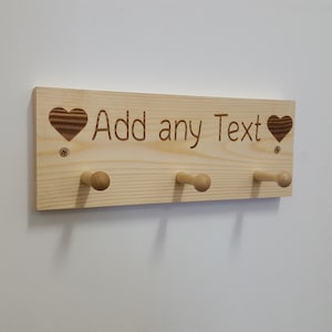 May include: A wooden coat rack with three hooks and two hearts carved into the wood. The text "Add any Text" is engraved on the rack.