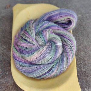 May include: A skein of yarn in a variety of colors, including purple, pink, green, and white. The yarn is sitting in a wooden bowl.