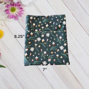 Puede incluir: Un artículo de tela verde oscuro con un estampado floral. El artículo mide 21 cm de largo y 18 cm de ancho. El estampado floral incluye flores blancas, naranjas y rosas con tallos y hojas verdes. El fondo es de color verde oscuro.
