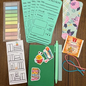 May include: A selection of stationery items for book enthusiasts, including sticky notes, book review cards, stickers, a green notebook, pen, highlighter, bookmark, tea bag, and elastic bands. The items are arranged on a wooden surface.