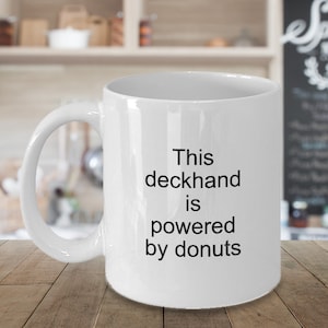 May include: White ceramic mug with a large handle. The mug features the text "This deckhand is powered by donuts" in black, centered on the front. The mug is set against a blurred background.