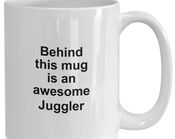 May include: White ceramic mug with a curved handle. The mug has the text "Behind this mug is an awesome Juggler" printed in black, centered on the front. The mug is designed for hot or cold beverages.