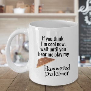 May include: White ceramic mug with the text "If you think I'm cool now, wait until you hear me play my Hammered Dulcimer" and a graphic of a dulcimer. The mug has a curved handle and is set against a blurred background.