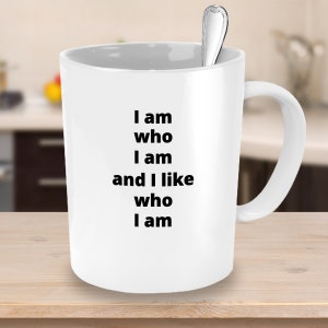 Puede incluir: Taza de cer&aacute;mica blanca con el texto "I am who I am and I like who I am" en negro. Una cuchara plateada descansa dentro de la taza. La taza tiene un asa curva y est&aacute; sobre un fondo de cocina borroso.