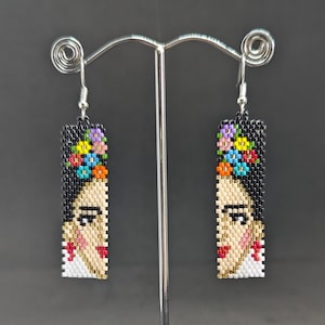 Peut inclure: Une paire de boucles d'oreilles en perles représentant un portrait de Frida Kahlo avec une couronne de fleurs. Les boucles d'oreilles sont noires, blanches, rouges, dorées et multicolores.