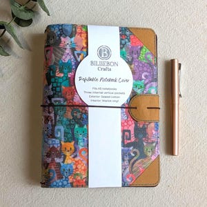 May include: A colorful refillable notebook cover with a cat pattern, a brown pen, and a white label. The notebook cover fits A5 notebooks and has three internal vertical pockets. The exterior is sealed cotton, and the interior is Warwick vinyl.