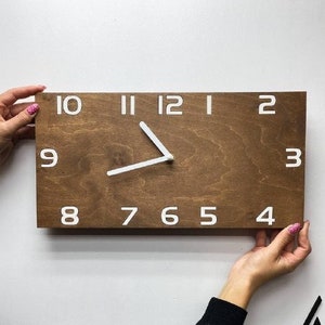 May include: A rectangular wooden wall clock with a dark brown finish. The clock face features white numerals and hands. The clock is being held up against a white wall.
