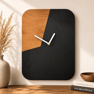 Op de afbeelding: Een rechthoekige wandklok met afgeronde hoeken met een gespleten ontwerp van natuurlijk lichtbruin houtnerf aan de linkerkant en massief matzwart hout aan de rechterkant, voorzien van twee eenvoudige witte wijzers in het midden.