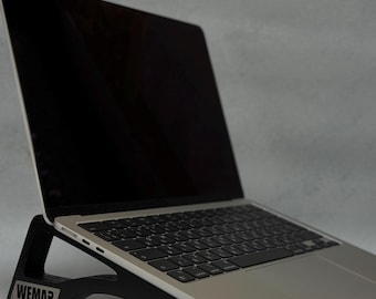 May include: A silver laptop computer is placed on a black laptop stand. The stand has the word "WEMAR" printed on the front. The laptop screen is black, and the keyboard is visible. The stand elevates the laptop.