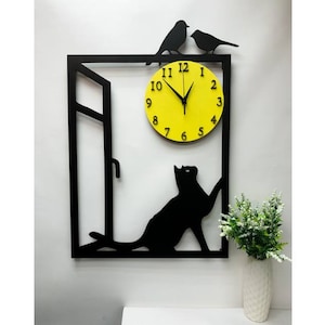 May include: Black metal wall clock with a yellow clock face and black numbers. The clock is shaped like a window with a cat silhouette looking out and two birds perched on top.