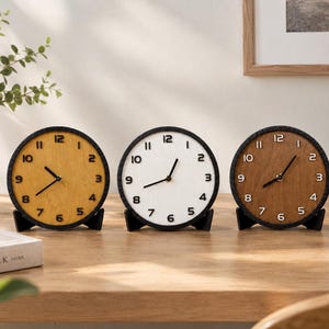 May include: Three circular desk clocks with black frames and stands. The left clock has a yellow face, the center clock has a white face, and the right clock has a brown face. Each clock features black numerals from one to twelve.