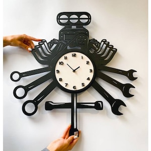 Puede incluir: Reloj de pared negro con diseño de motor de coche. La esfera del reloj es blanca con números y manecillas negros. El motor es de metal negro y tiene un diseño de llave inglesa a cada lado.