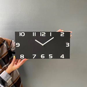 Puede incluir: Un reloj de pared rectangular negro con números y manecillas blancas. La esfera del reloj muestra los números del 1 al 12 en una fuente moderna. Las manecillas son blancas e indican la hora. El reloj se sostiene sobre un fondo gris.