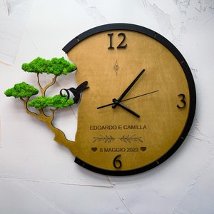 May include: A wooden clock with a black frame and a green moss tree design. The clock face has black hands and Roman numerals. The clock also has the text "Edoardo e Camilla" and "6 Maggio 2023" on it.
