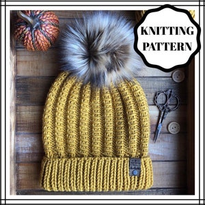 Puede incluir: Un gorro de punto amarillo mostaza con un pompón de piel sintética. El gorro tiene una textura acanalada y un pequeño parche de cuero marrón con el texto "NORTHSTAR" en él. El gorro está sobre una superficie de madera con unas tijeras y botones.