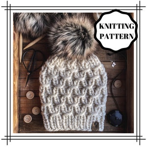 May include: A white knitted beanie with a fluffy brown and black pom pom. The beanie has a textured stitch pattern and a brown leather tag that says "Knitting Pattern".