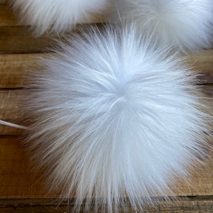 May include: Three white fluffy pom poms, made of faux fur, are arranged on a wooden surface. The pom poms are soft and plush, and they would be perfect for adding a touch of whimsy to any project.
