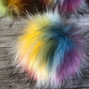 May include: A multi-colored fluffy pom pom with shades of yellow, blue, white, and purple. The pom pom is made of faux fur and is a popular accessory for hats, scarves, and other winter wear.