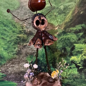 May include: A handcrafted fairy ornament made from natural elements. The fairy features an acorn head, a walnut shell body, and a skirt of dried leaves. It is mounted on a wooden base decorated with small flowers and foliage.
