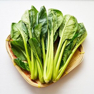 Op de afbeelding: Een mandje gevuld met verse groene bladgroenten, waarschijnlijk Aziatische groenten, op een witte achtergrond. De bladeren zijn heldergroen, met lichtgroene stelen. De mand is gemaakt van lichtbruin geweven materiaal.