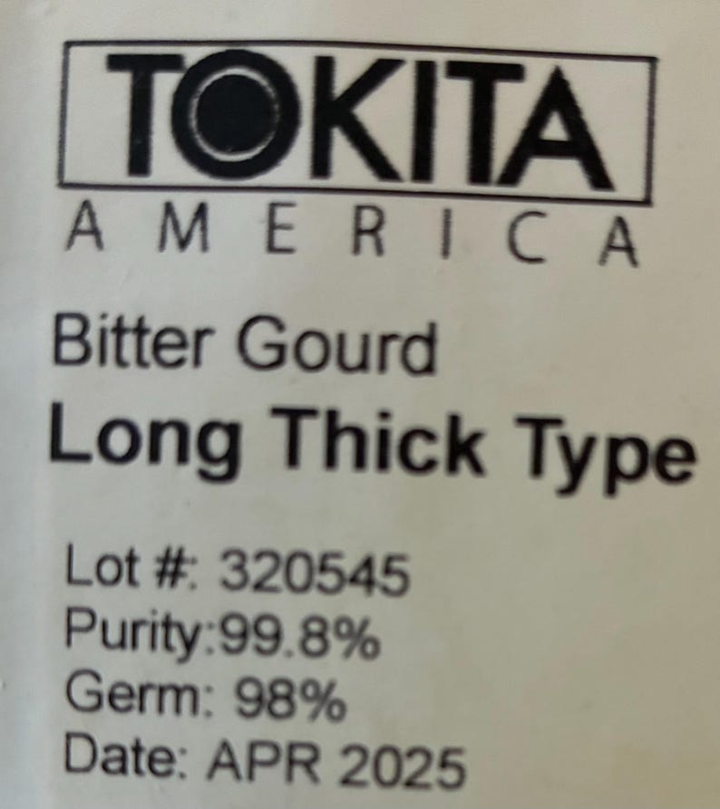 May include: A white label with black text. The text reads "TOKITA AMERICA", "Bitter Gourd Long Thick Type", Lot # 320545, Purity: 99.8%, Germ: 98%, and Date: APR 2025. The label is for a food product.