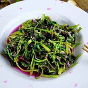 May include: A white plate with a serving of stir-fried red amaranth greens. The greens are a vibrant green and red color and are arranged in a pile on the plate. The plate has pink splatter paint on it.