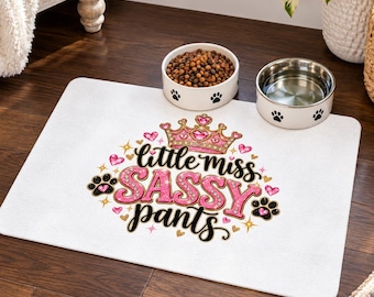 May include: A rectangular white pet feeding mat featuring a graphic design with a pink glittery crown, pink hearts, black paw prints, and the text "little miss SASSY pants" in black and pink glittery script lettering.
