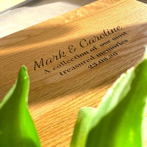 Pode incluir: Uma t&aacute;bua de corte de madeira com texto gravado. O texto diz "Mark & Caroline, A collection of our most treasured memories, 23.08.86". A t&aacute;bua &eacute; castanho-claro com gr&atilde;o de madeira natural.