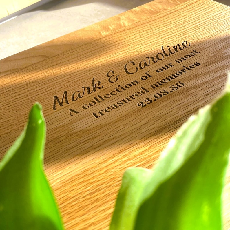 Pode incluir: Uma t&aacute;bua de corte de madeira com texto gravado. O texto diz "Mark & Caroline, A collection of our most treasured memories, 23.08.86". A t&aacute;bua &eacute; castanho-claro com gr&atilde;o de madeira natural.