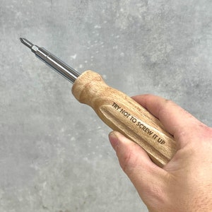 May include: A screwdriver with a light-colored wooden handle and a silver metal shaft. The handle features the engraved text "TRY NOT TO SCREW IT UP" in a dark font. The tool has a standard Phillips head tip at the end of the metal shaft.