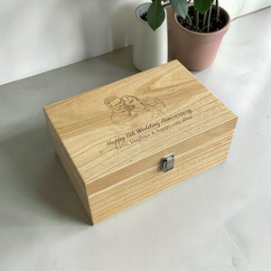Peut inclure: Une boîte à souvenirs rectangulaire en bois clair avec un fermoir en métal argenté. Le couvercle présente un dessin au trait gravé au laser d'un couple s'enlaçant et le texte : Happy 6th Wedding Anniversary, Love, laughter & happy ever after.