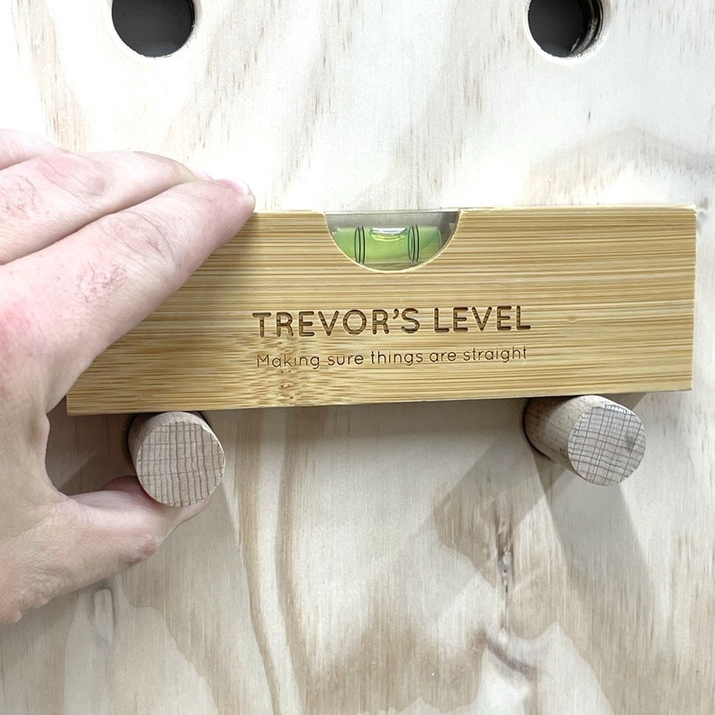 Pu&ograve; includere: Un livello in legno con un livello a bolla verde e il testo "TREVOR'S LEVEL Making sure things are straight" inciso su di esso.
