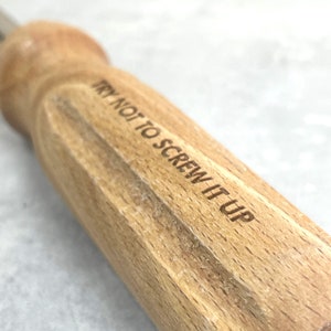 May include: A wooden screwdriver handle featuring the laser-engraved text TRY NOT TO SCREW IT UP. The handle has a light brown natural wood grain finish with a rounded top and a cylindrical shape, showing clear wood texture and grain patterns.