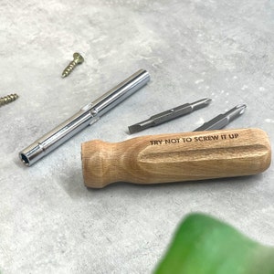 May include: A screwdriver set featuring a light brown wooden handle engraved with the text TRY NOT TO SCREW IT UP, a silver metal shaft, and two interchangeable double-ended screwdriver bits, accompanied by two small metal screws.